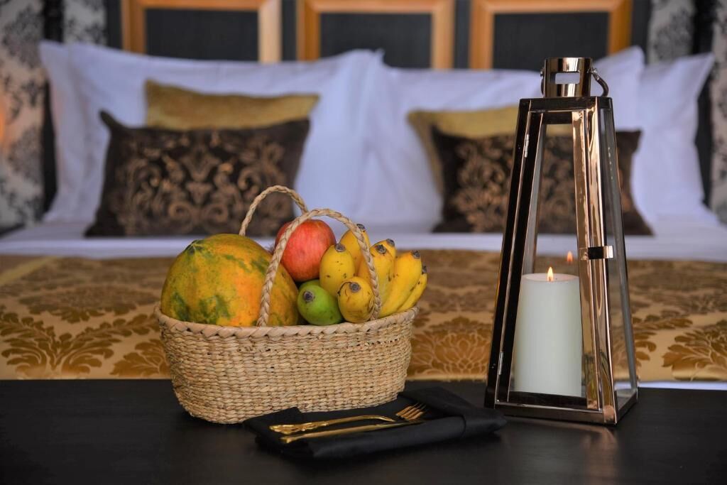 Un lit recouvert d’une couvre-lit blanche et dorée, un panier en osier rempli de bananes et de mangues, une bougie allumée dans une lanterne en métal, et une serviette noire avec une fourchette dorée, le tout posé sur une table.