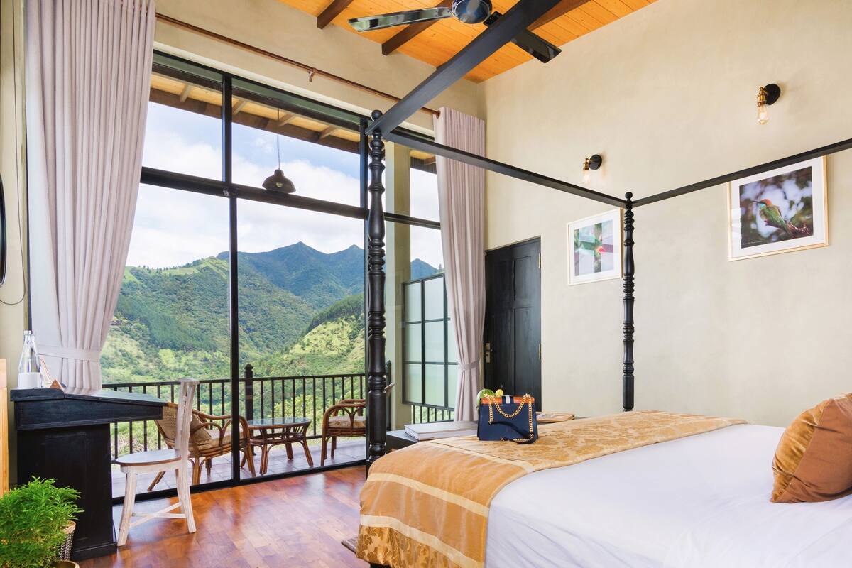 A bedroom with a large bed, a desk, and a chair. There is a view of mountains through large windows.