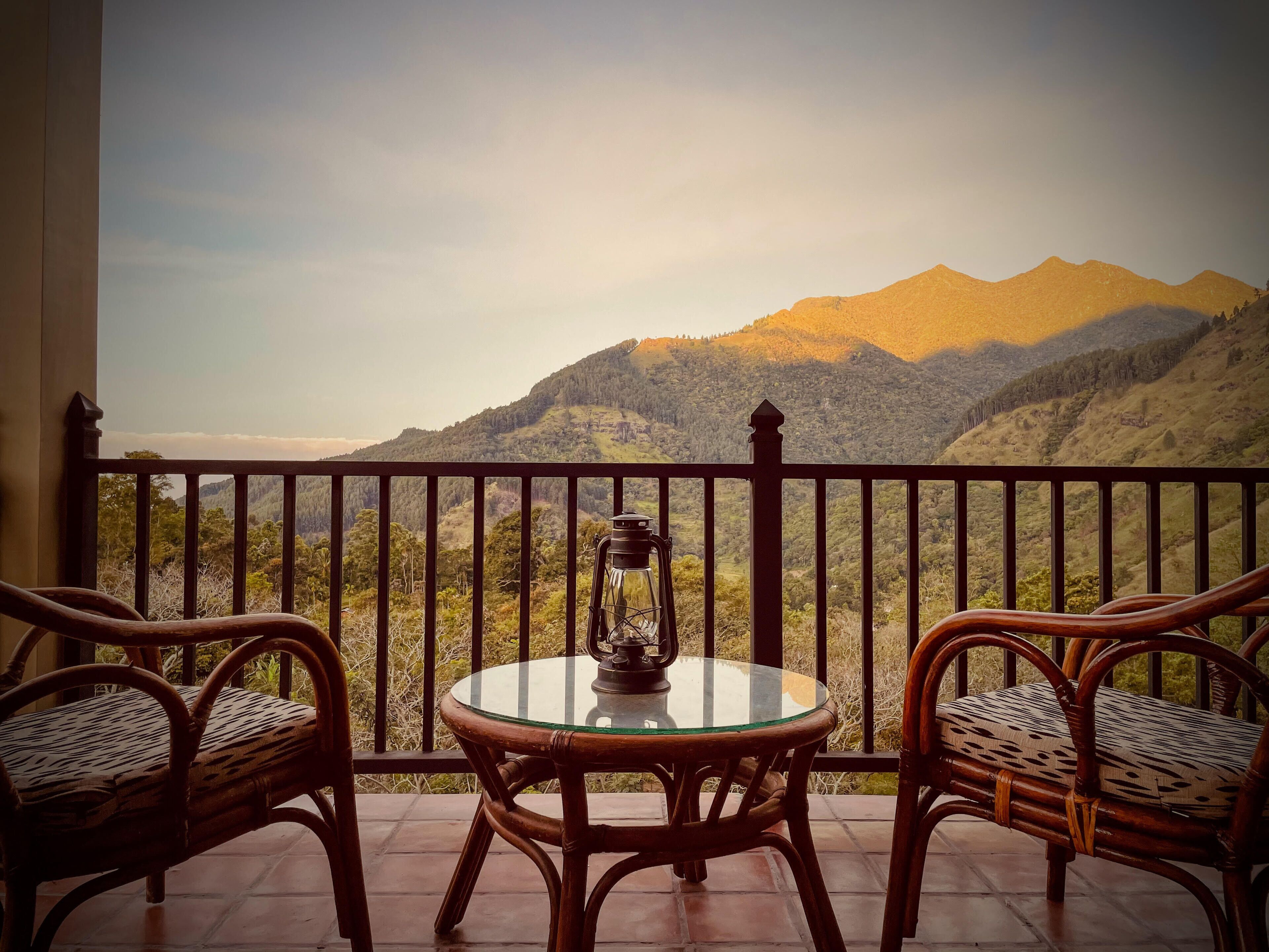 A balcony with wicker chairs and a glass table, overlooking a mountainous landscape.