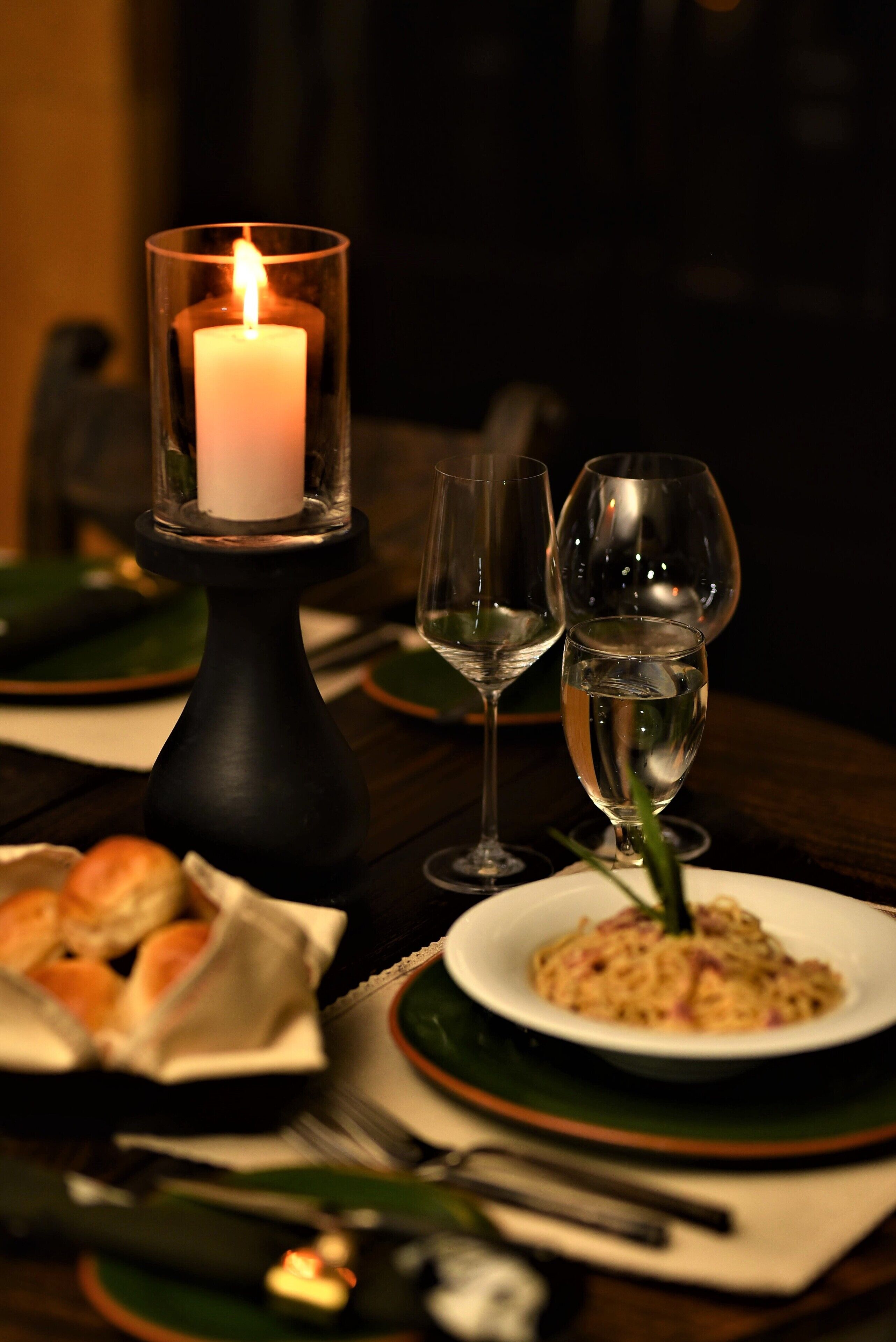 Een kaars in een glazen houder op een tafel met een bord eten en twee wijnglazen.