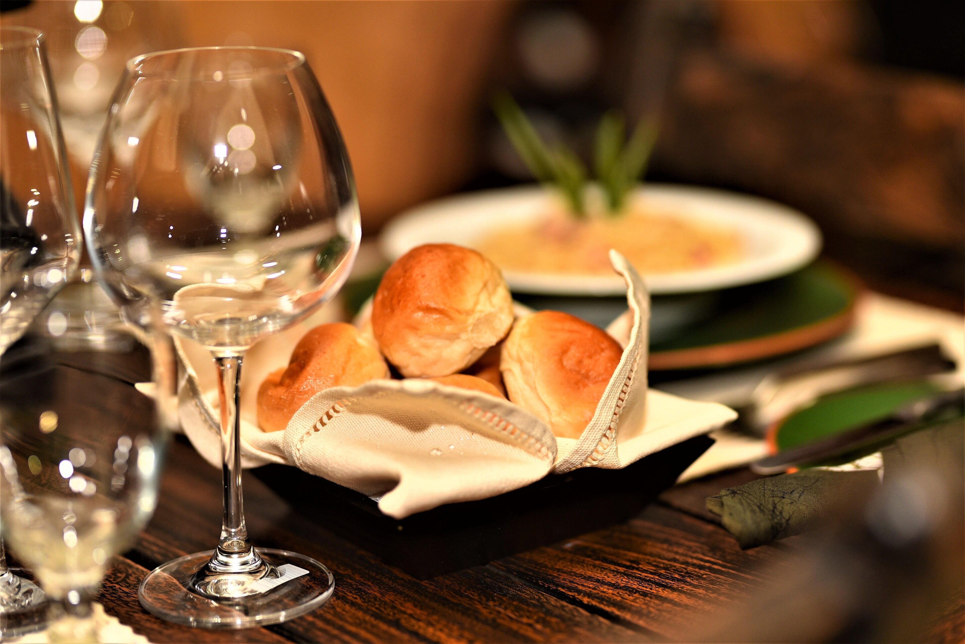 A dining table with wine glasses, a basket of bread rolls, and a plate of soup.