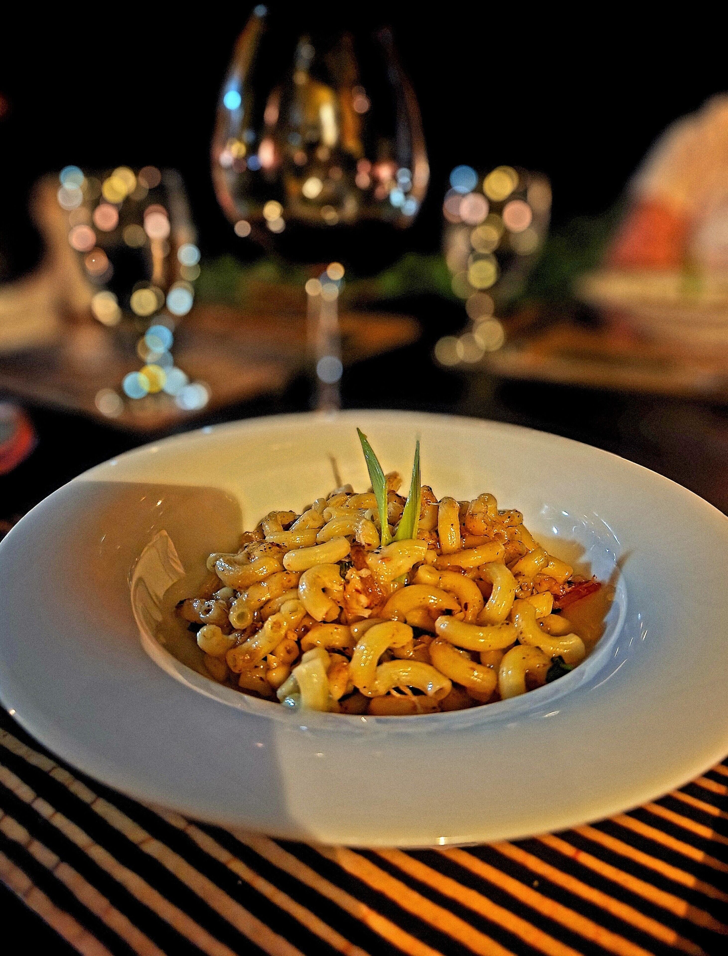 A white plate with a pasta dish, a glass of wine, and a blurred background with lights.