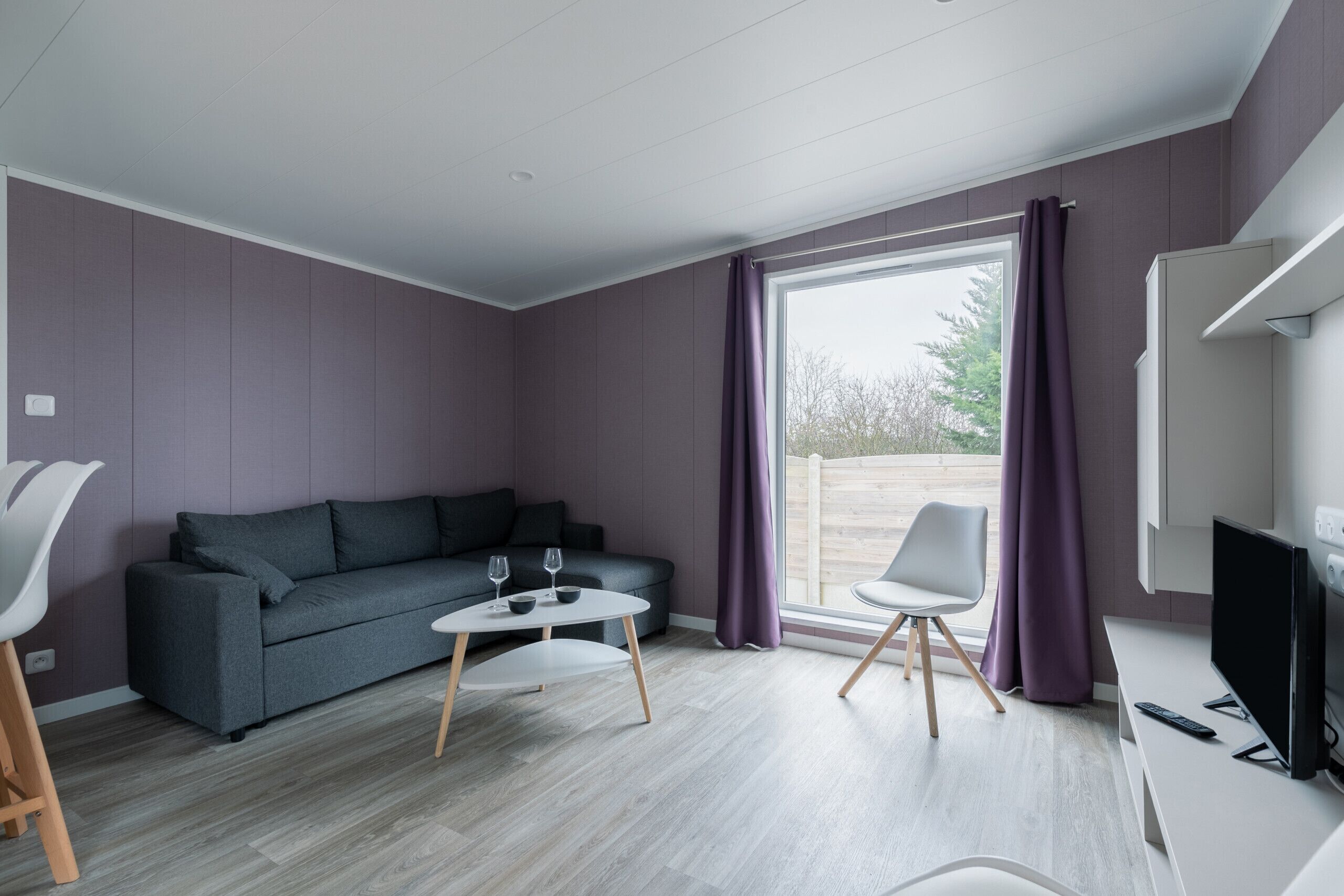 A modern living room with a grey sofa, a small round table, two chairs, a TV, and a large window with purple curtains.