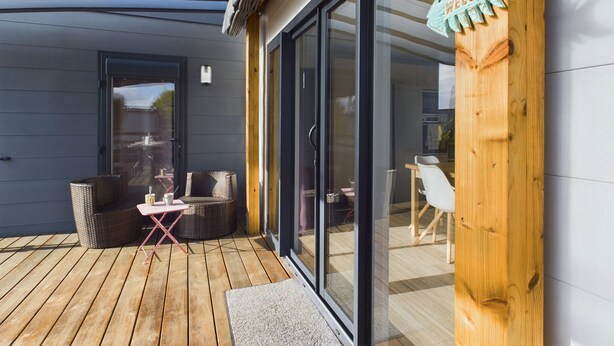 A modern building with a wooden deck, wicker furniture, and a glass door. - Beautiful Chalet for 4 people - Heated pool access (Bretignolles-sur-Mer)