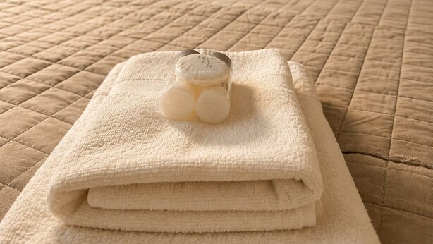 Un lit bien fait, recouvert d’une courtepointe matelassée, une pile de serviettes pliées et un petit flacon de lotion posé sur une serviette.