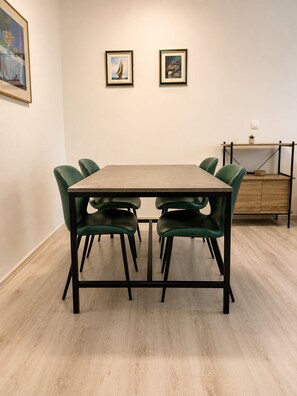 A dining area with a rectangular table, six green chairs, two framed artworks on the wall, and a wooden cabinet with a plant.