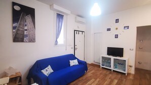 A living room with a blue sofa, a television on a white cabinet, a framed picture on the wall, and a window with checkered curtains. - B&BO - IN BOLOGNA YOU DREAM - APARTMENT - FREE PARKING  (BOLOGNA)