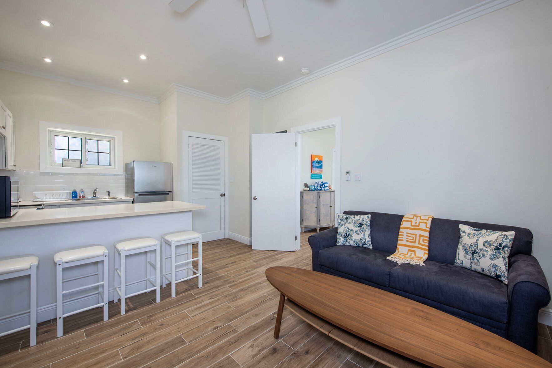 A compact living space with a kitchenette, a dark blue sofa with patterned cushions, a wooden bench, and a white door leading to another room.