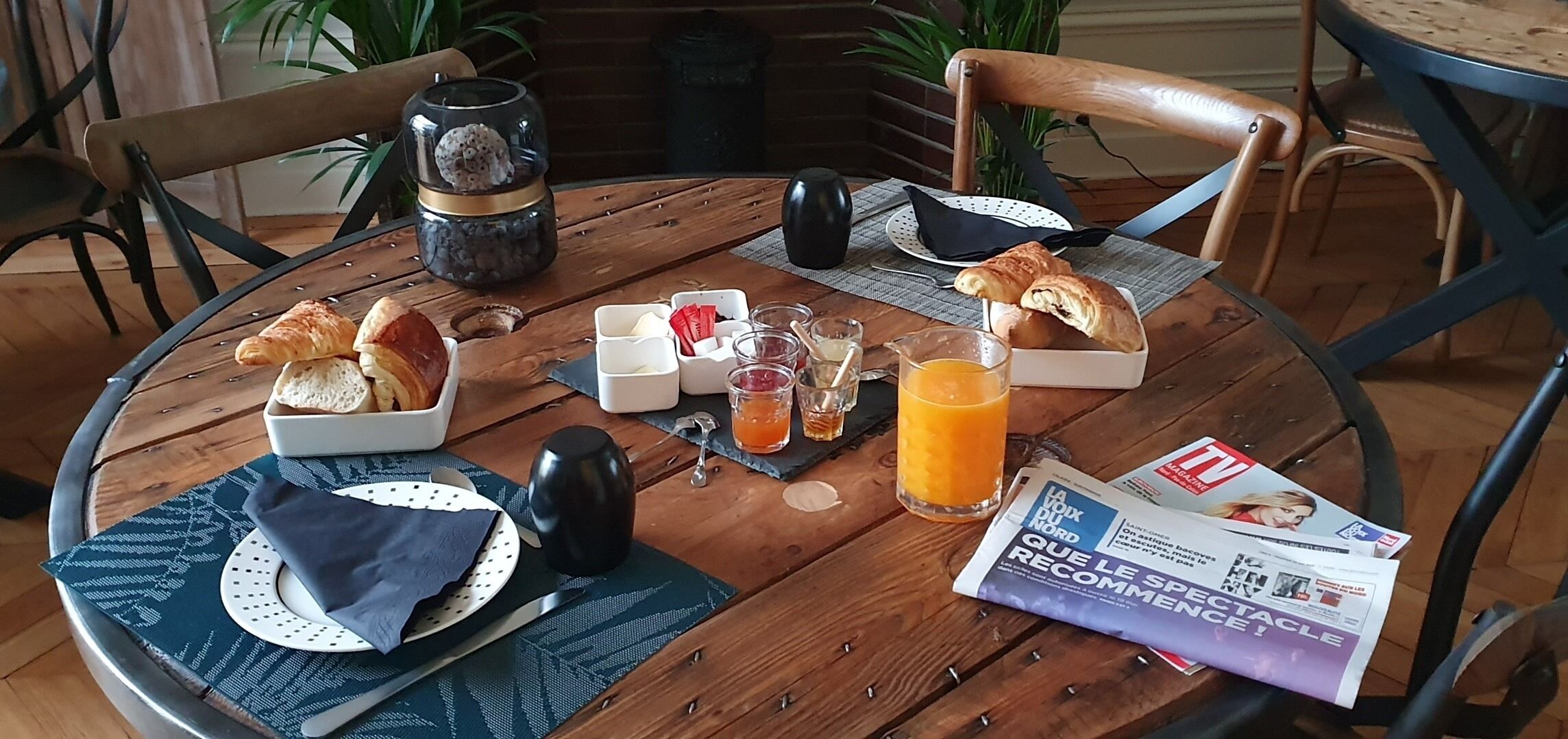Une table ronde en bois, sur laquelle sont disposés des aliments pour le petit-déjeuner, notamment des croissants, du fromage et du jus, ainsi qu’un journal et un vase décoratif.