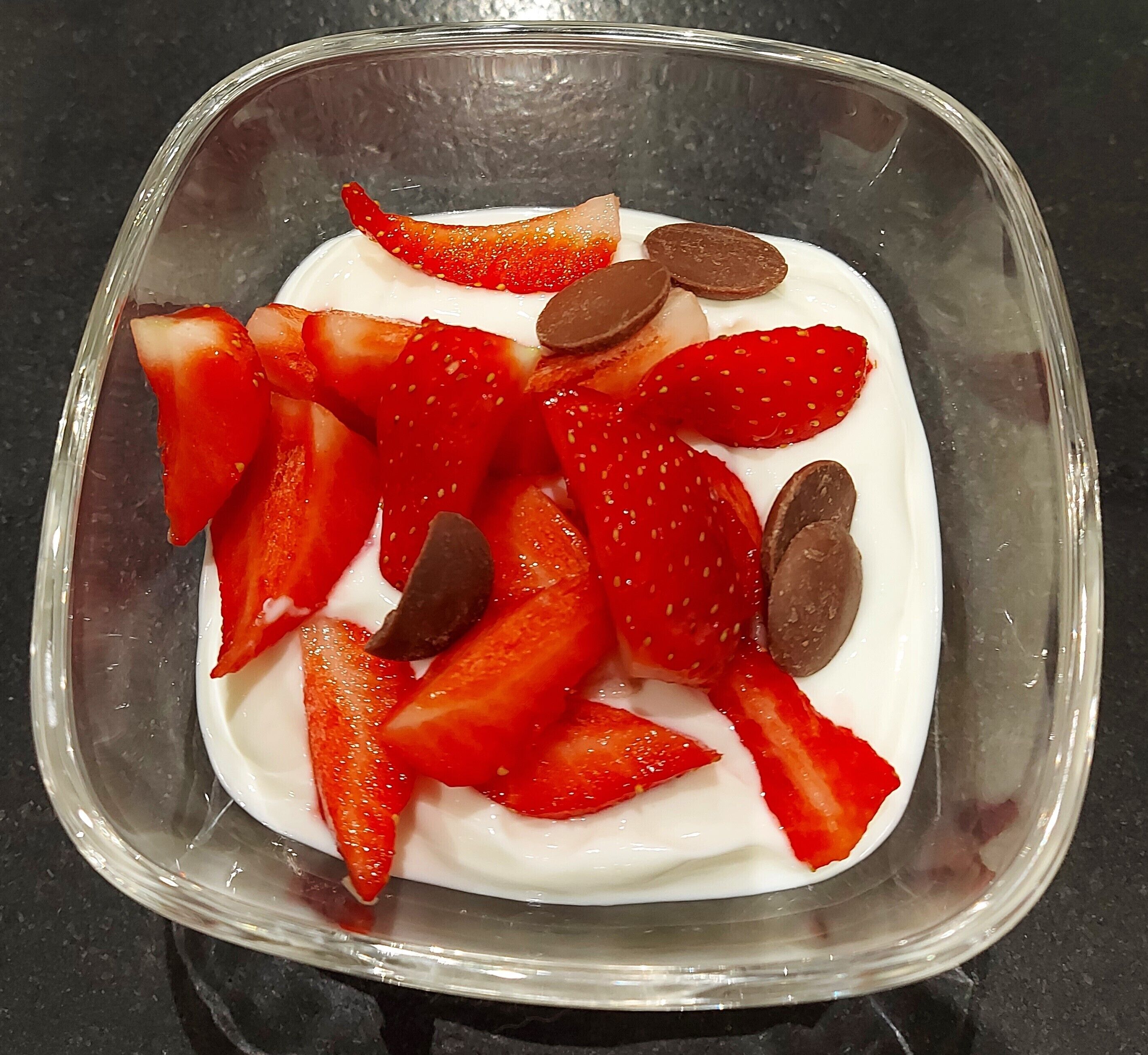 Een glazen schaal met plakjes aardbeien en stukjes chocolade op een witte ondergrond.