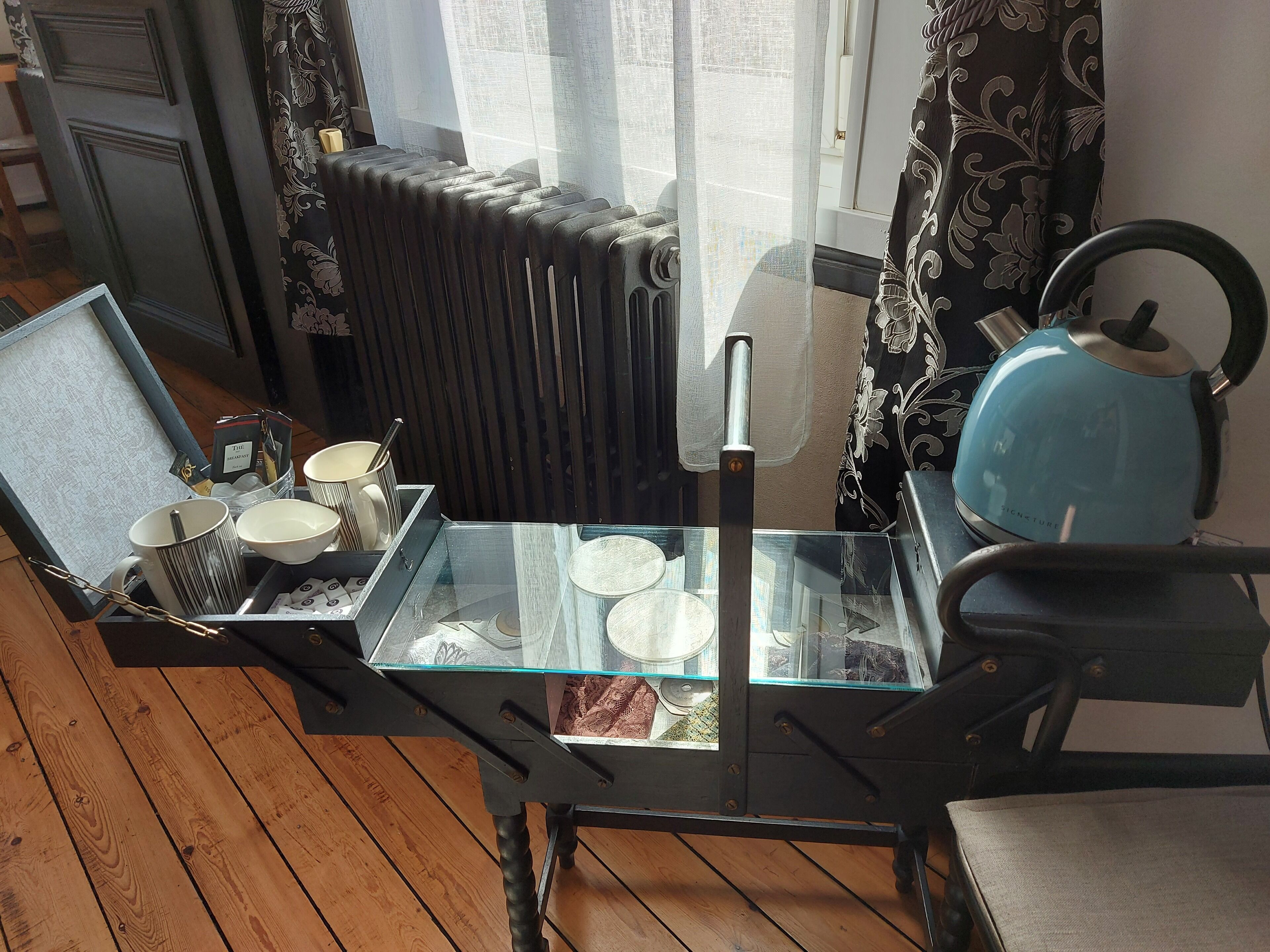 A dining table with a glass top, surrounded by chairs, in a room with a radiator and patterned curtains.