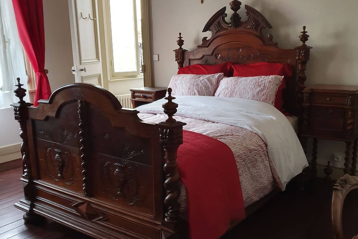 A traditional wooden four-poster bed with a red and white bedspread, a nightstand with a lamp, and a red curtain on the window.