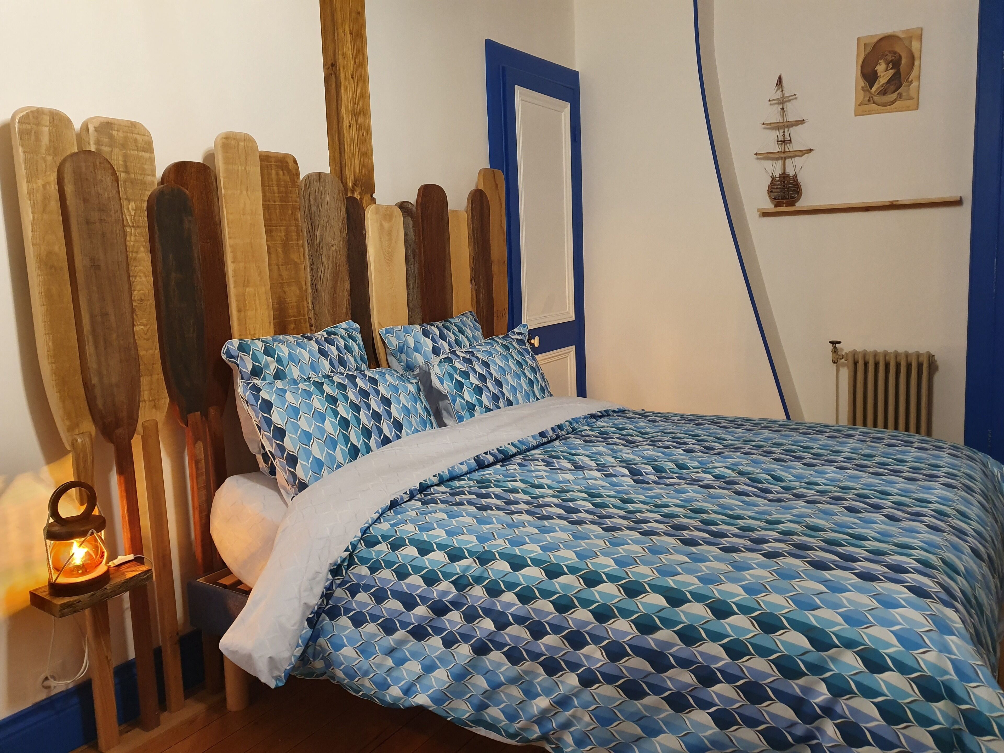 Une chambre à coucher avec une tête de lit en bois, un lit recouvert d’une courtepointe à motifs bleus, et une petite étagère sur laquelle sont posés un modèle de bateau et un portrait.