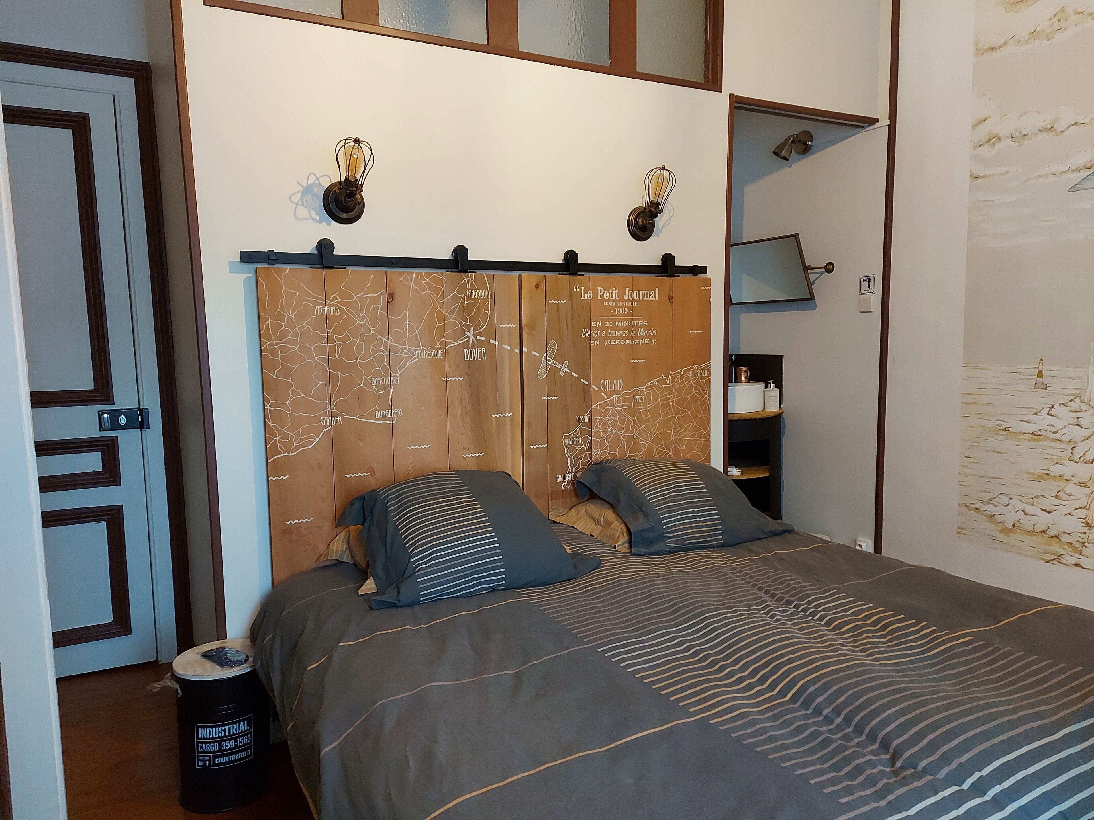 Een slaapkamer met een bed, een houten hoofdbord met een kaart, een nachtkastje en een prullenbak.