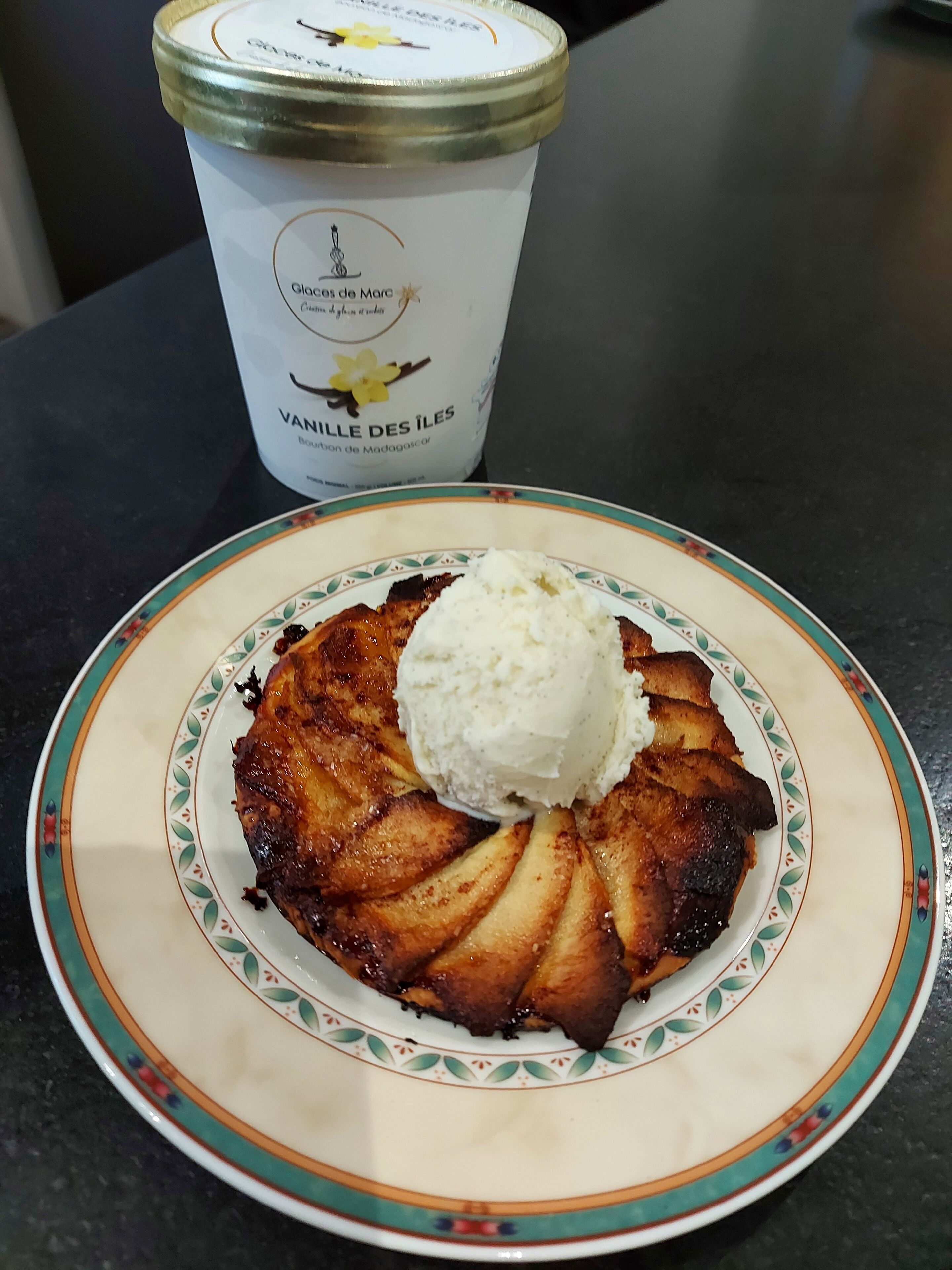 A plate with a caramelized apple dessert and a scoop of vanilla ice cream, placed on a table with a white container labeled "VANILLE DES ÎLES."