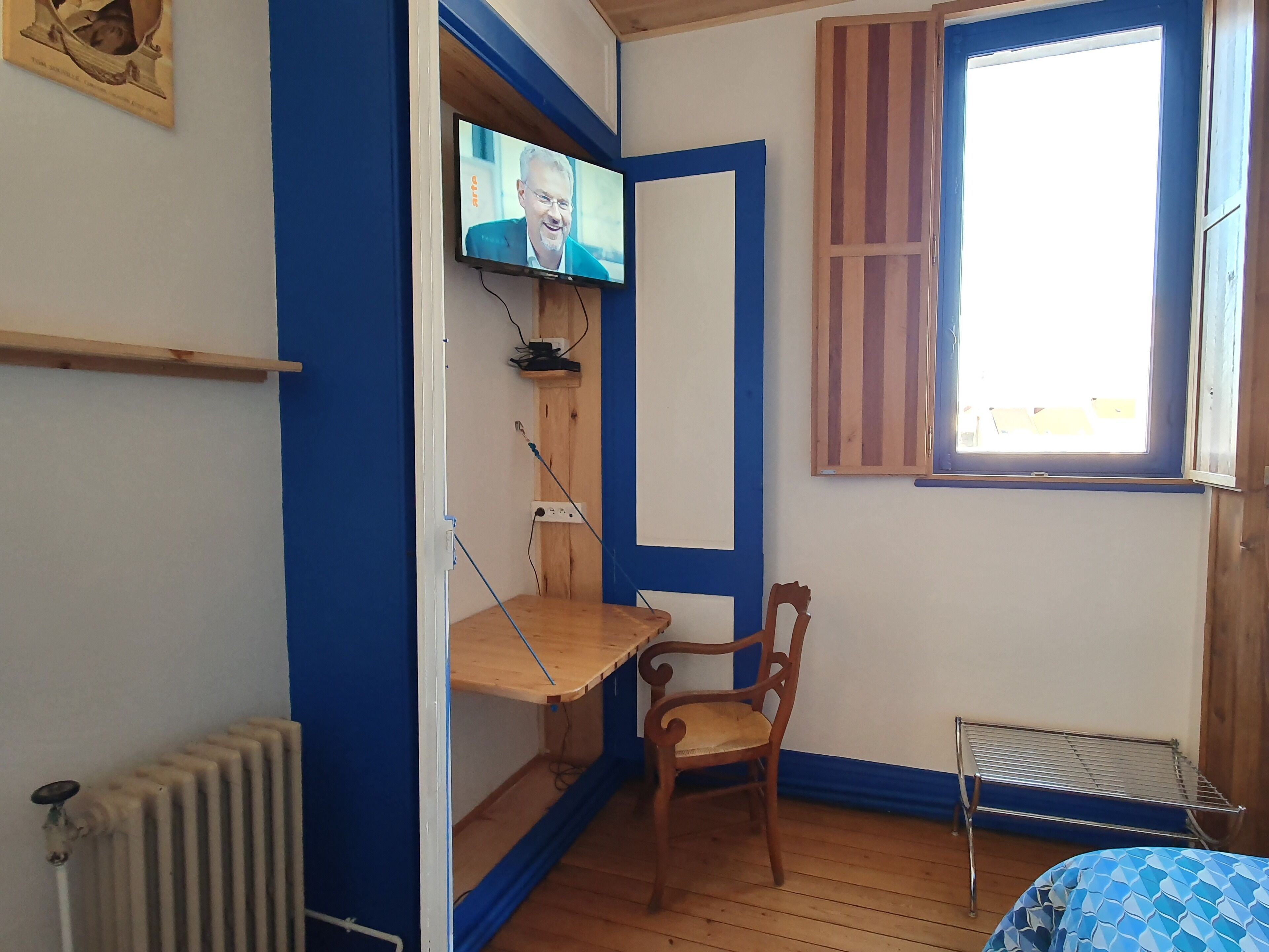 A room with a wooden desk, a chair, a radiator, a television mounted on the wall, and a window with wooden shutters.