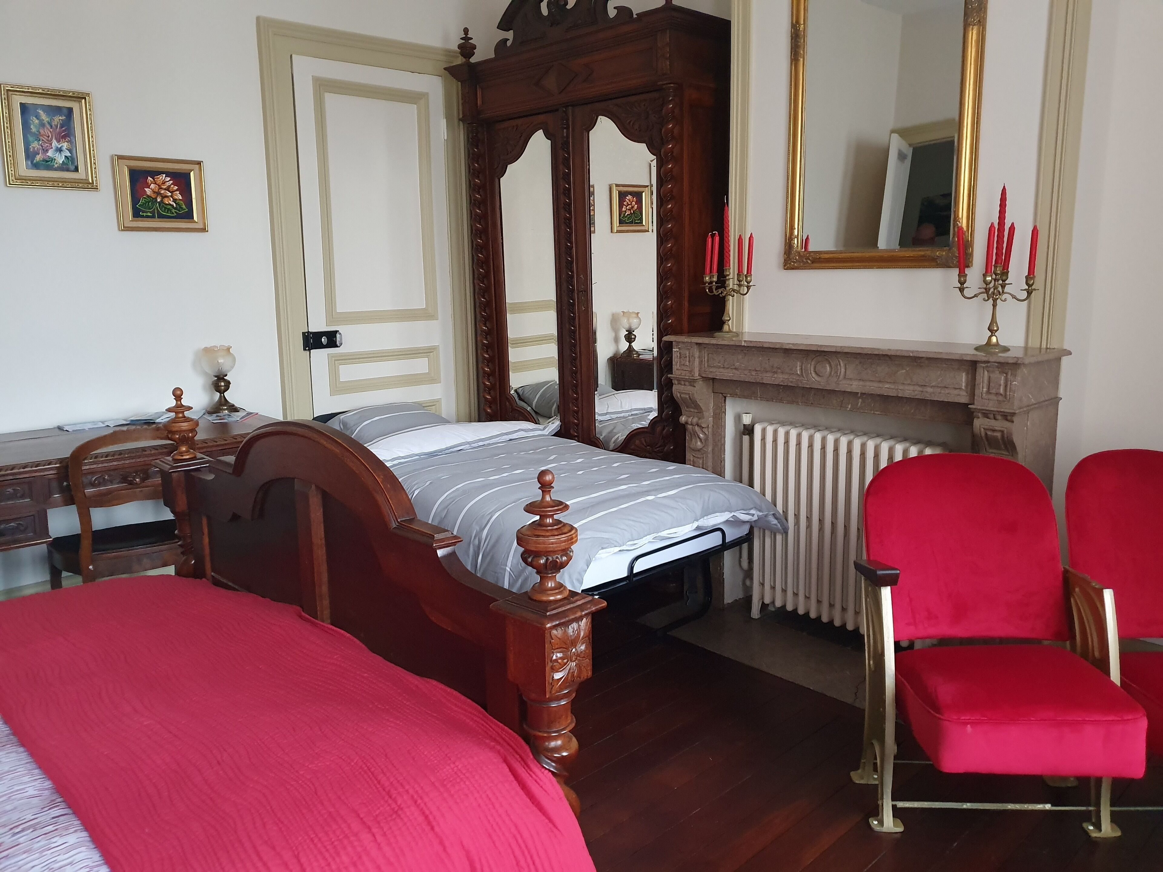 A bedroom with a four-poster bed, a red chair, a wooden dresser, and a mirror.