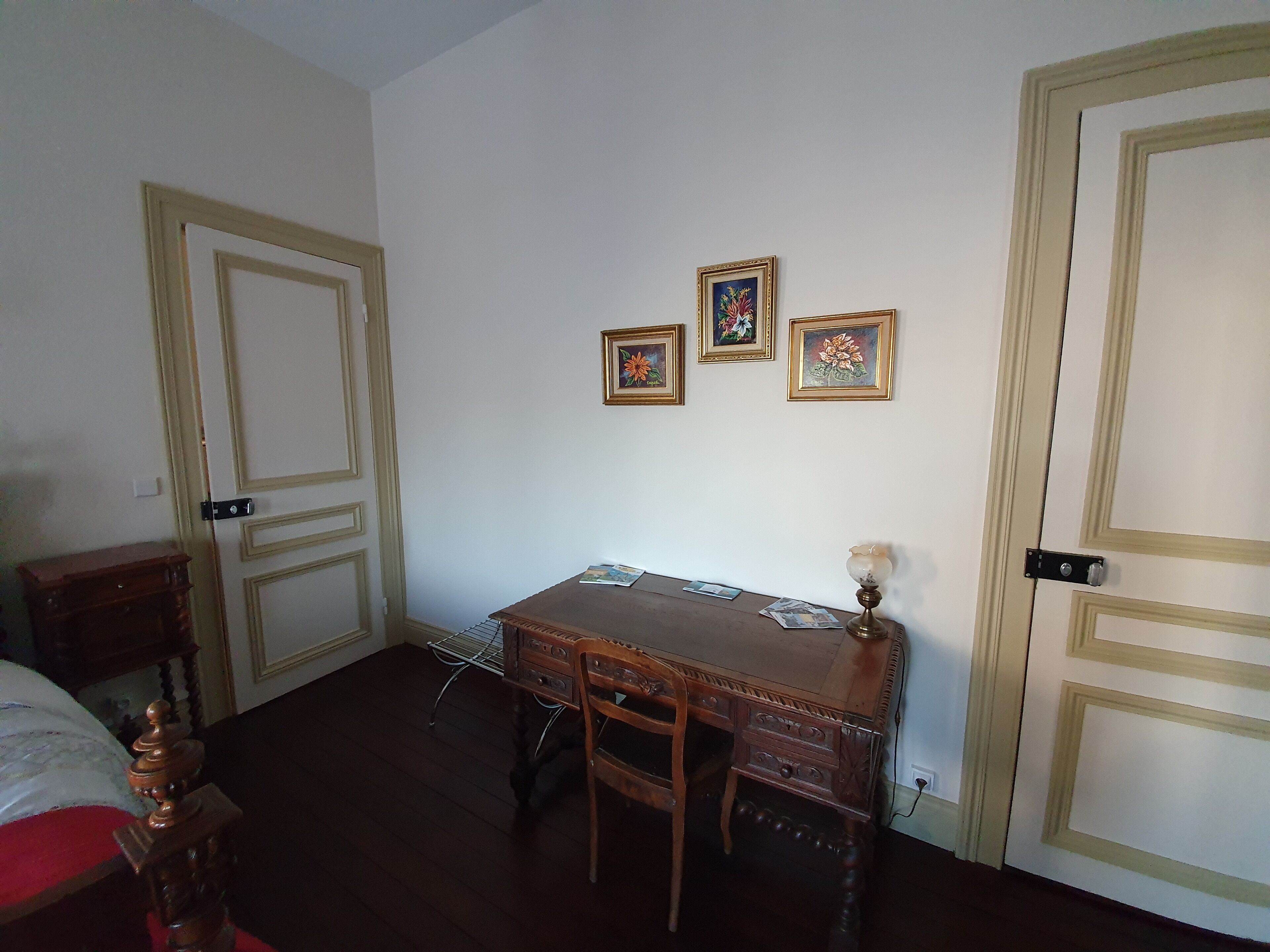 A room with a wooden desk, chairs, and a lamp. There are three framed pictures on the wall.