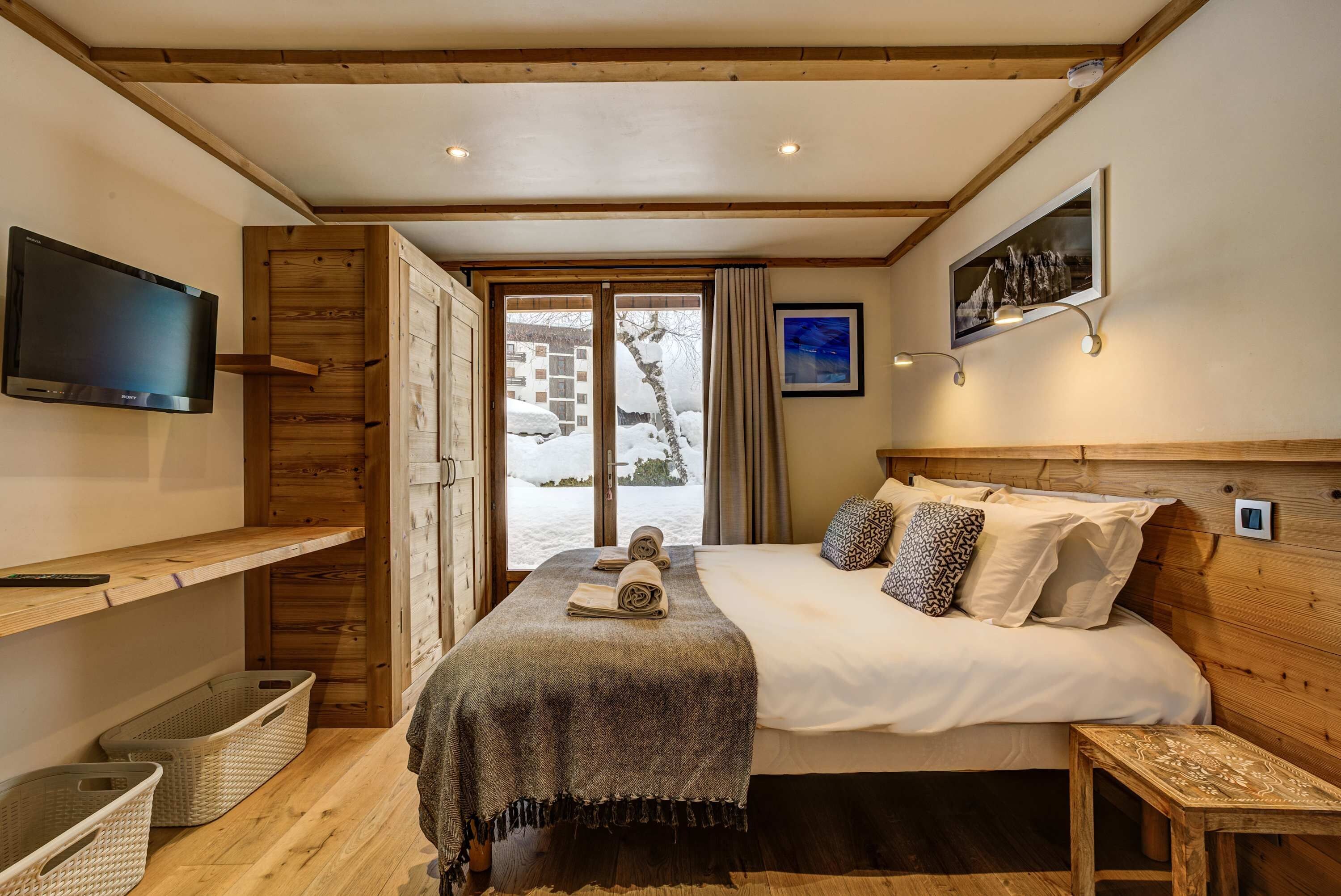 Une chambre d’hôtel avec un lit, une télévision, une fenêtre donnant sur des bâtiments enneigés et un plafond en bois.
