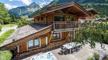 Un chalet in legno con vasca idromassaggio e area salotto esterna, circondato da montagne.