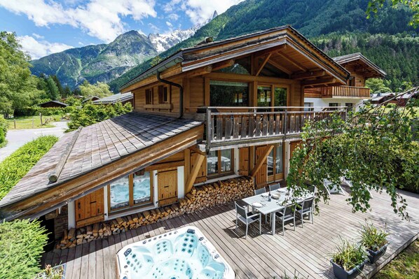 A wooden chalet with a hot tub and outdoor seating, surrounded by mountains.