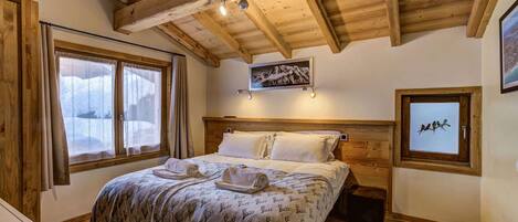 Une chambre à coucher avec un plafond en bois, un lit recouvert d’une couverture à motifs, deux lampes de chevet et un tableau encadré accroché au mur.