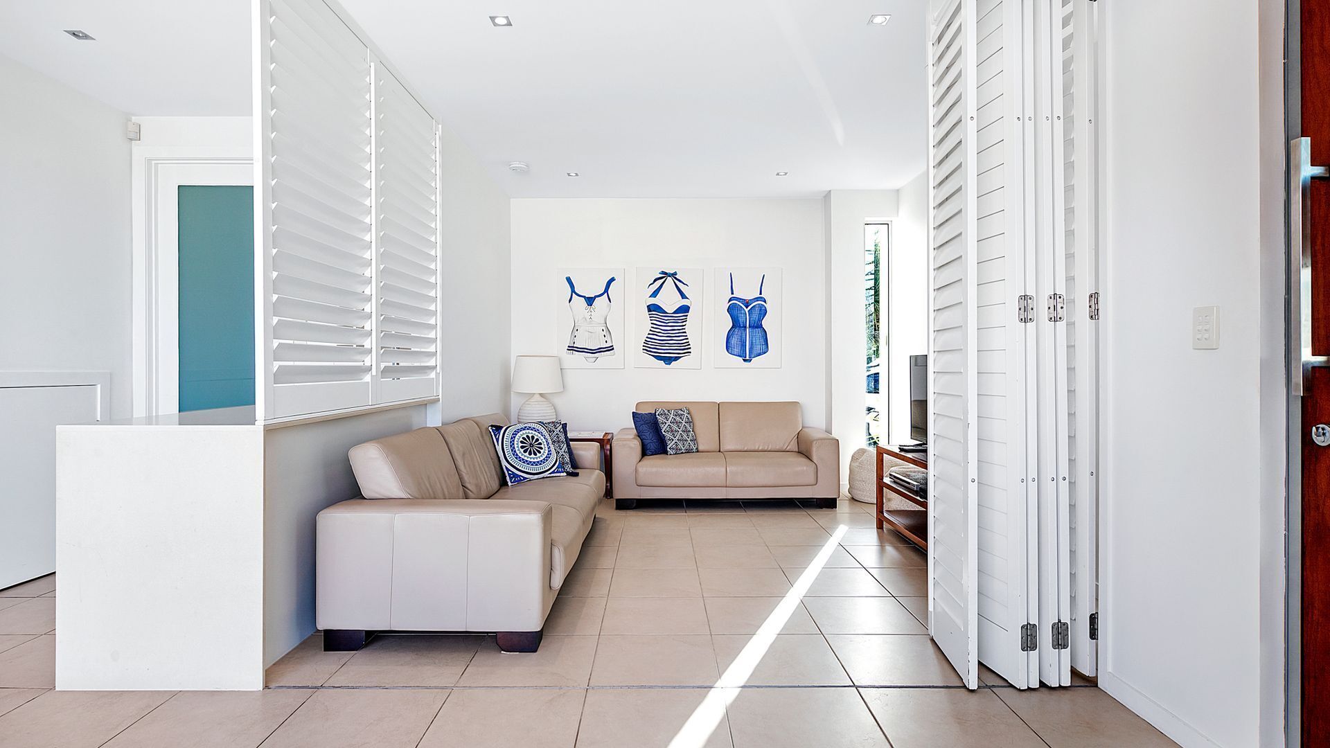 Una sala de estar moderna con un sofá color beige, una mesa de centro blanca y un cojín azul y blanco estampado. Se observan tres obras de arte enmarcadas en la pared, una puerta blanca y un armario blanco con un espejo.