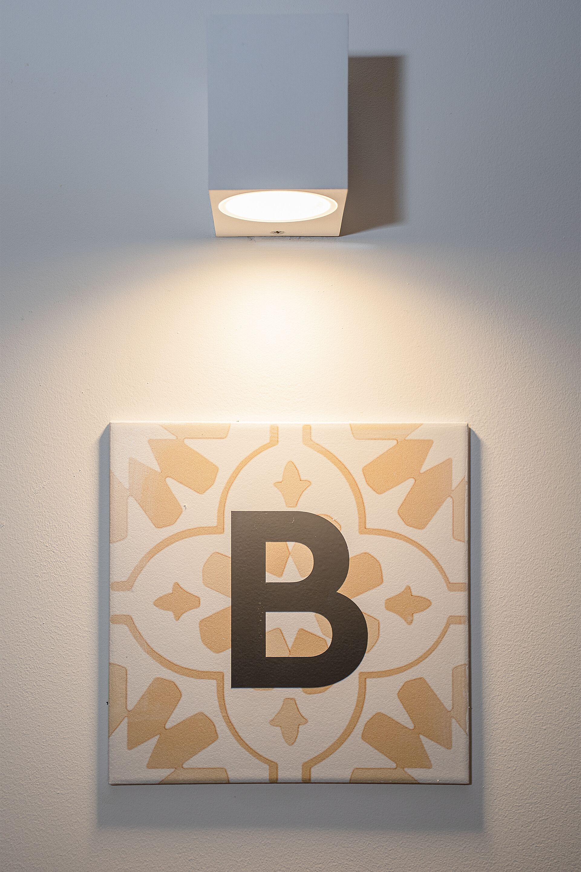 Un luminaire mural orné d’un motif géométrique, avec un grand panneau lumineux en forme de « B » placé en dessous.