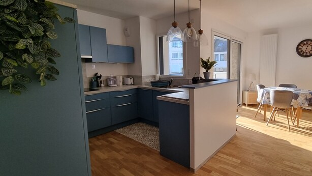 Een moderne keuken met blauwe kastjes, een eethoek met een tafel en stoelen, en een groot raam dat voor natuurlijk licht zorgt.