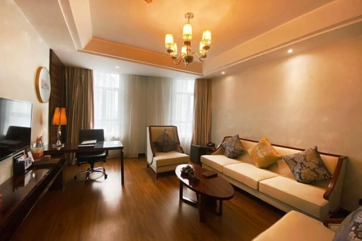Ein Hotelzimmer mit Sofa, Sesseln, Schreibtisch, Fernseher und einer Chandelier.