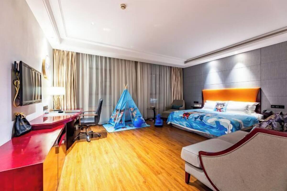 Ein Hotelzimmer mit Holzboden, einem Schreibtisch mit roter Oberfläche, einem Bett mit blau-weißer, an das Meer erinnernden Bettdecke und einem großen Fenster mit Vorhängen.