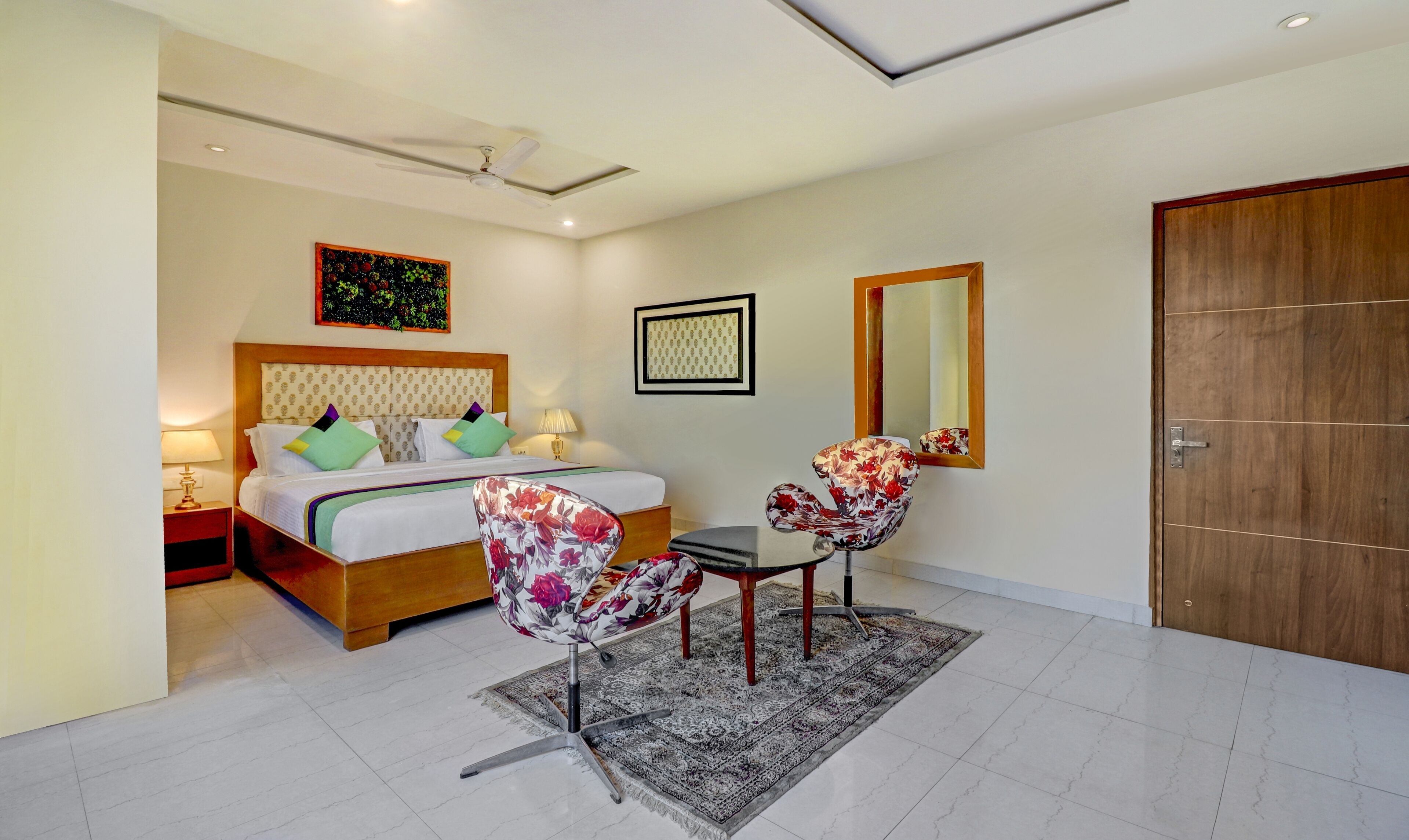 A hotel room with a bed, two chairs, a nightstand, a bedside table, a ceiling fan, a framed picture, a mirror, and a wooden door.