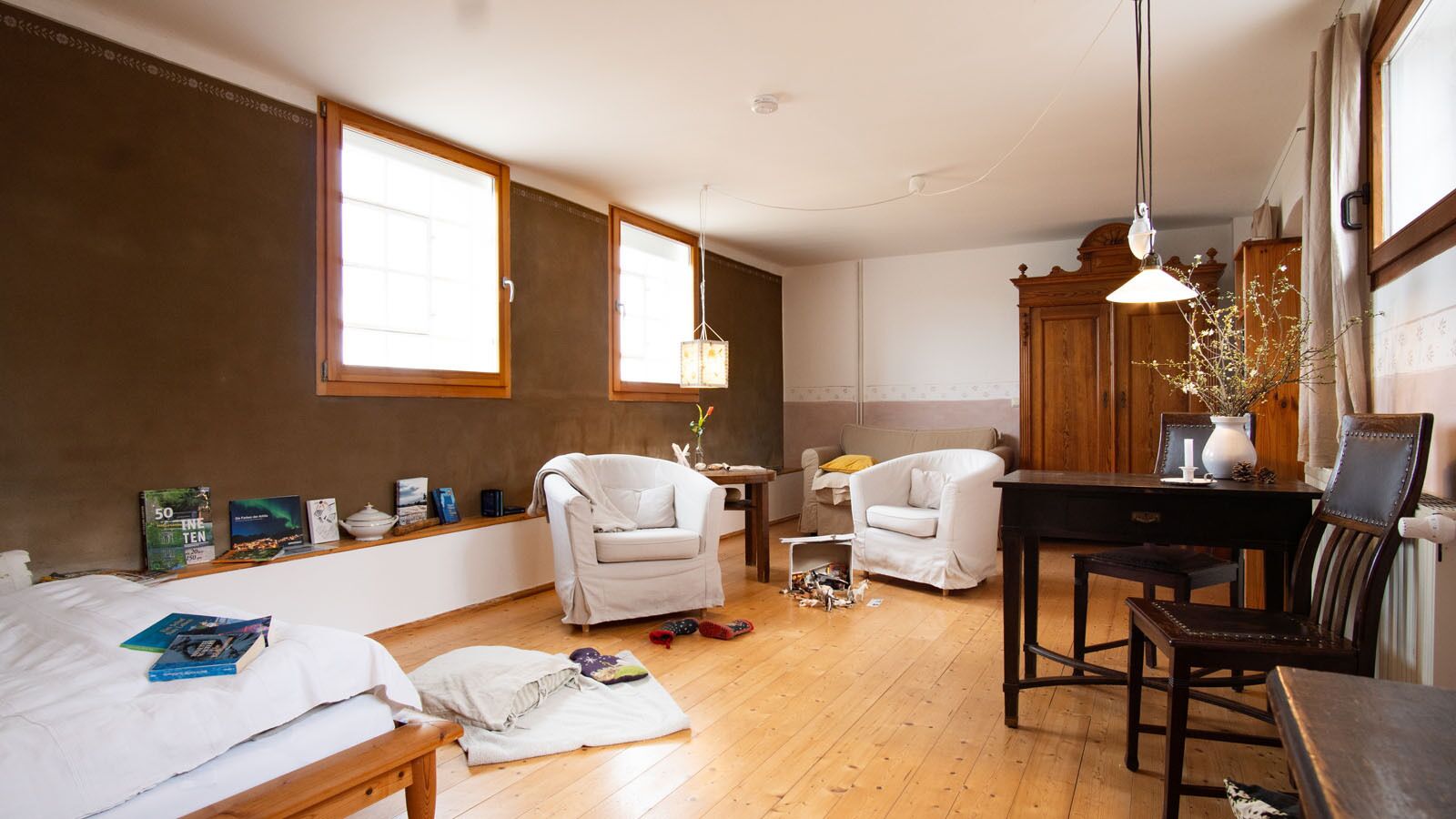 A room with a bed, two white armchairs, a wooden table, and a desk.
