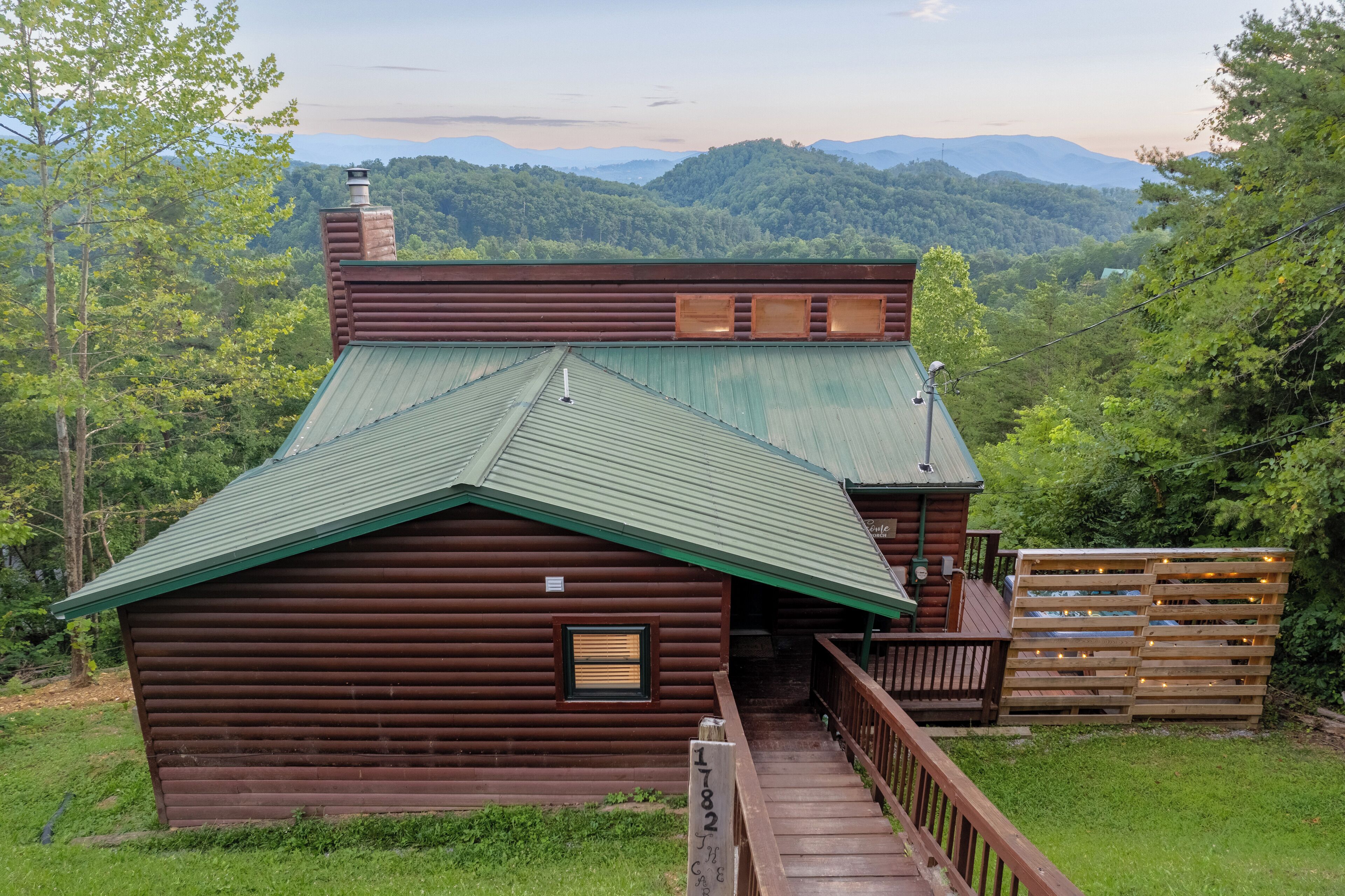 A rustic cabin with a green metal roof, wooden deck, and a view of a forested mountain range.