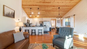 A spacious living area with a wooden ceiling, a brown leather sofa, a grey armchair, and a kitchen in the background. - STUNNING SMOKY MOUNTAIN VIEWS! Hot Tub! Games! Close to Dollywood and Parkway! (Sevierville)