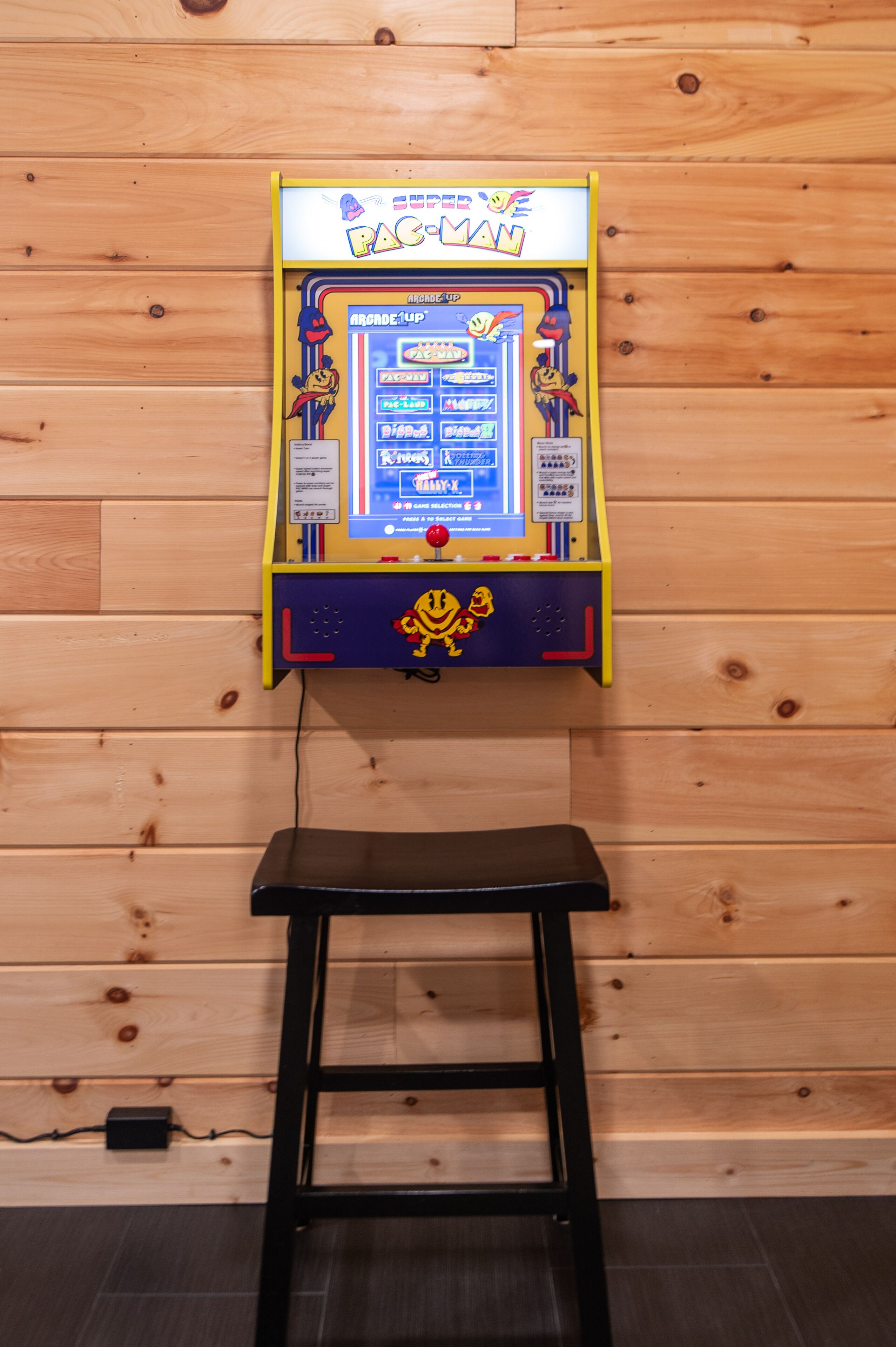 A Pac-Man arcade machine mounted on a wooden wall with a black stool in front.