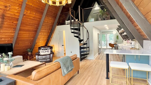 A loft-style living area with a wooden ceiling, a sofa, a dining table, and a staircase leading to an upper level. - Amazing A-Frame Cabin, Modern Luxury Oasis on a Pond (Pittsboro)