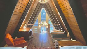 A cozy attic bedroom with a wooden sloped ceiling, a chandelier, a patterned rug, and a view of the outdoors through a large window. - Amazing A-Frame Cabin, Modern Luxury Oasis on a Pond (Pittsboro)