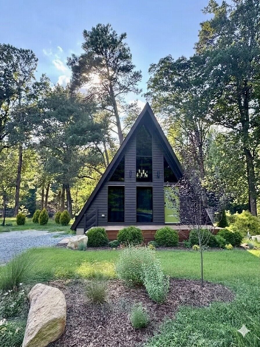 Un bâtiment moderne de forme triangulaire, doté d’une grande fenêtre et entouré d’un jardin bien entretenu, agrémenté de diverses plantes et d’un élément décoratif en roche.