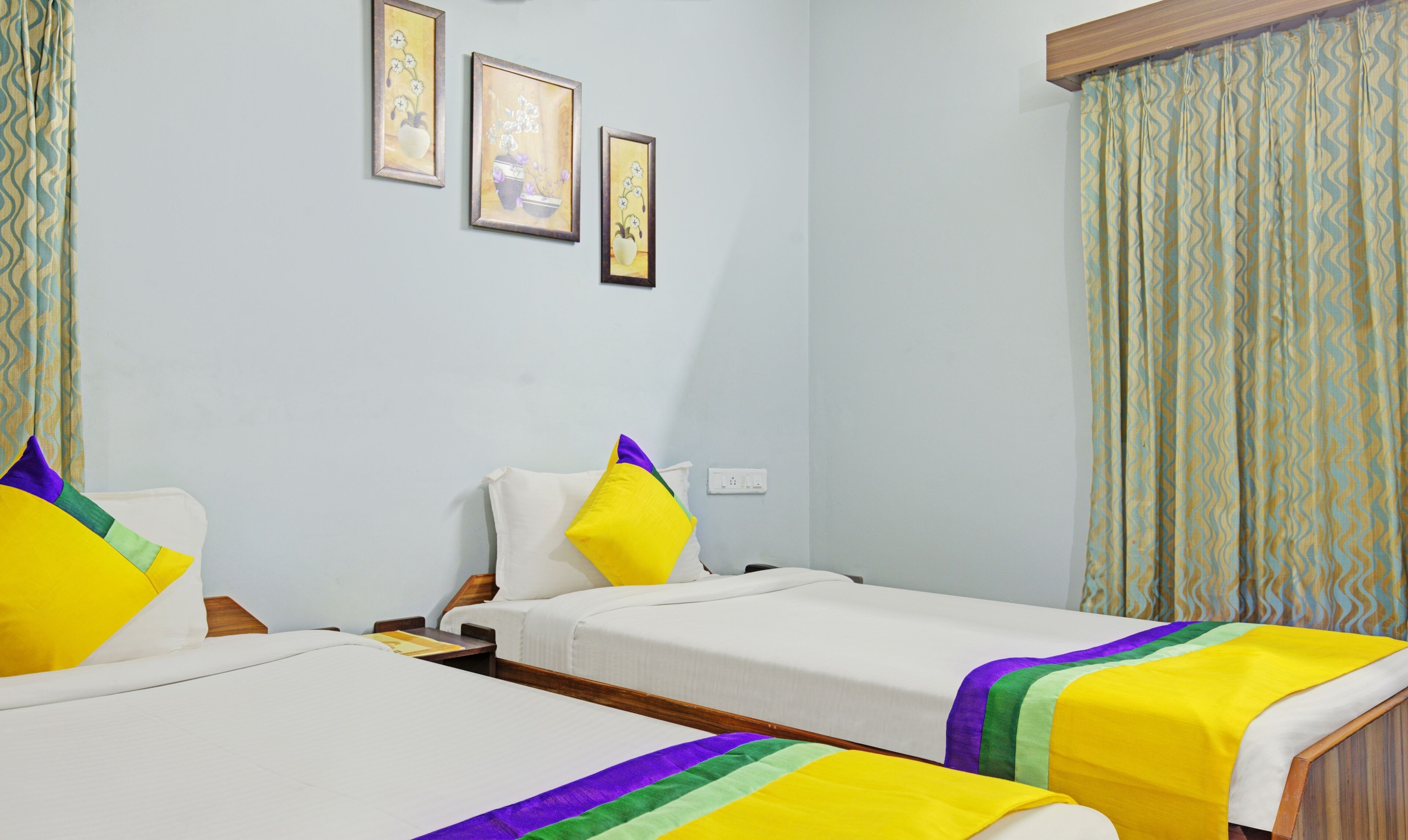 A hotel room with two beds, each with a white bedspread and colorful pillows. There are three framed pictures on the wall, and a window with patterned curtains.