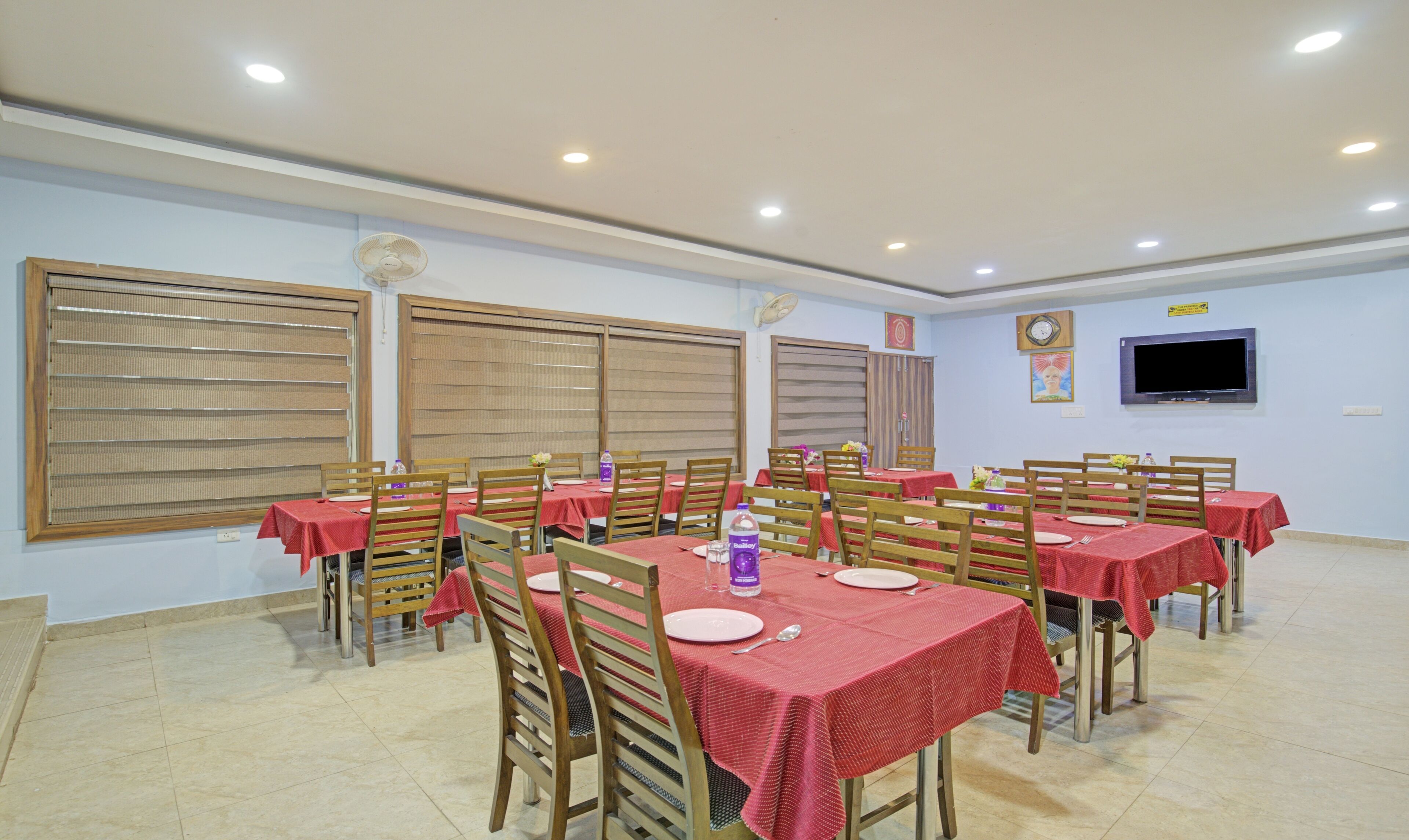 A dining area with multiple tables and chairs, equipped with ceiling lights and large windows with blinds.