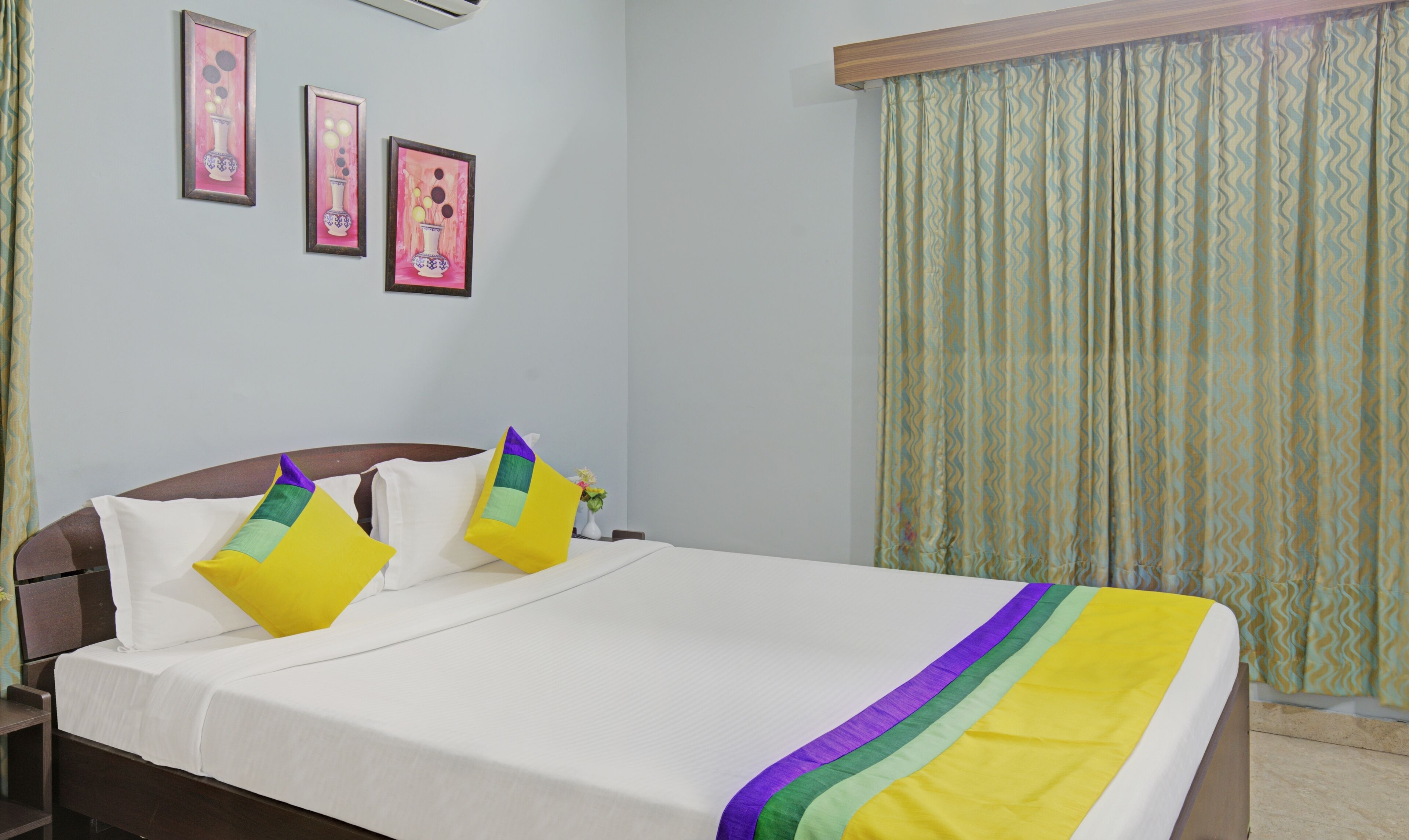 Una habitación de hotel con cama, almohadas, mesita de noche, un jarrón con flores y dos cuadros en la pared.