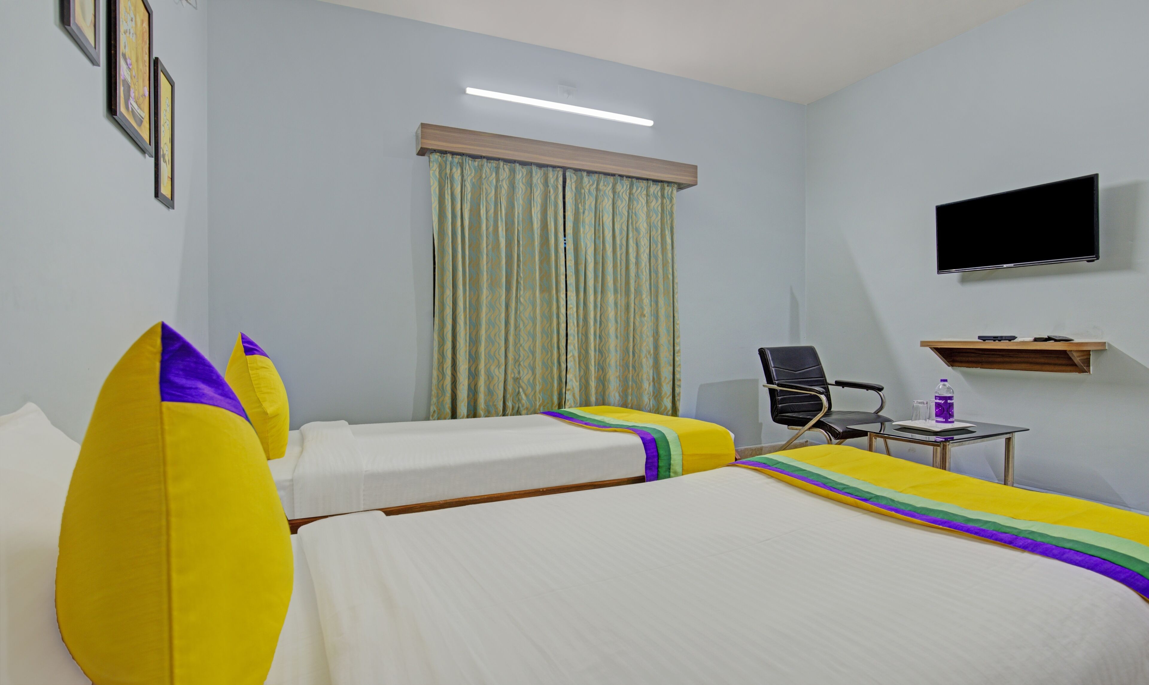 A hotel room with two single beds, a desk with a chair, a flat-screen TV, and a window with curtains.