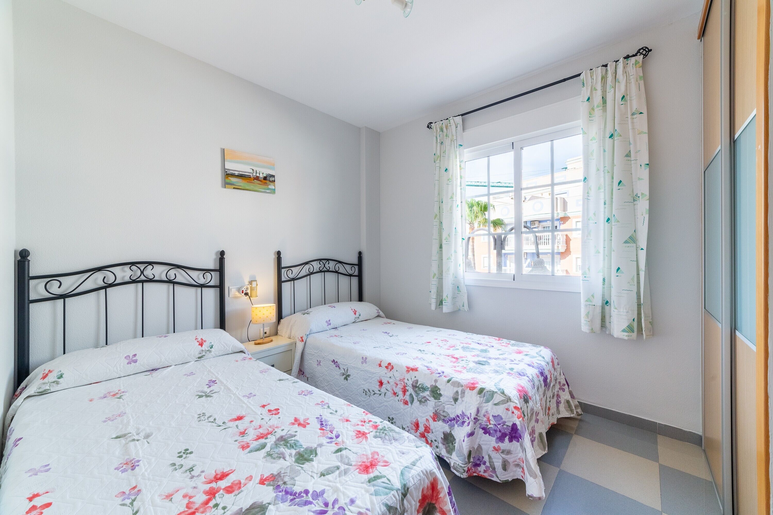 Un dormitorio con dos camas, una ventana con cortinas, una mesita de noche con lámpara y un cuadro colgado en la pared.