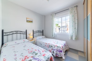 A bedroom with two beds, a window with curtains, a bedside table with a lamp, and a wall-mounted picture.