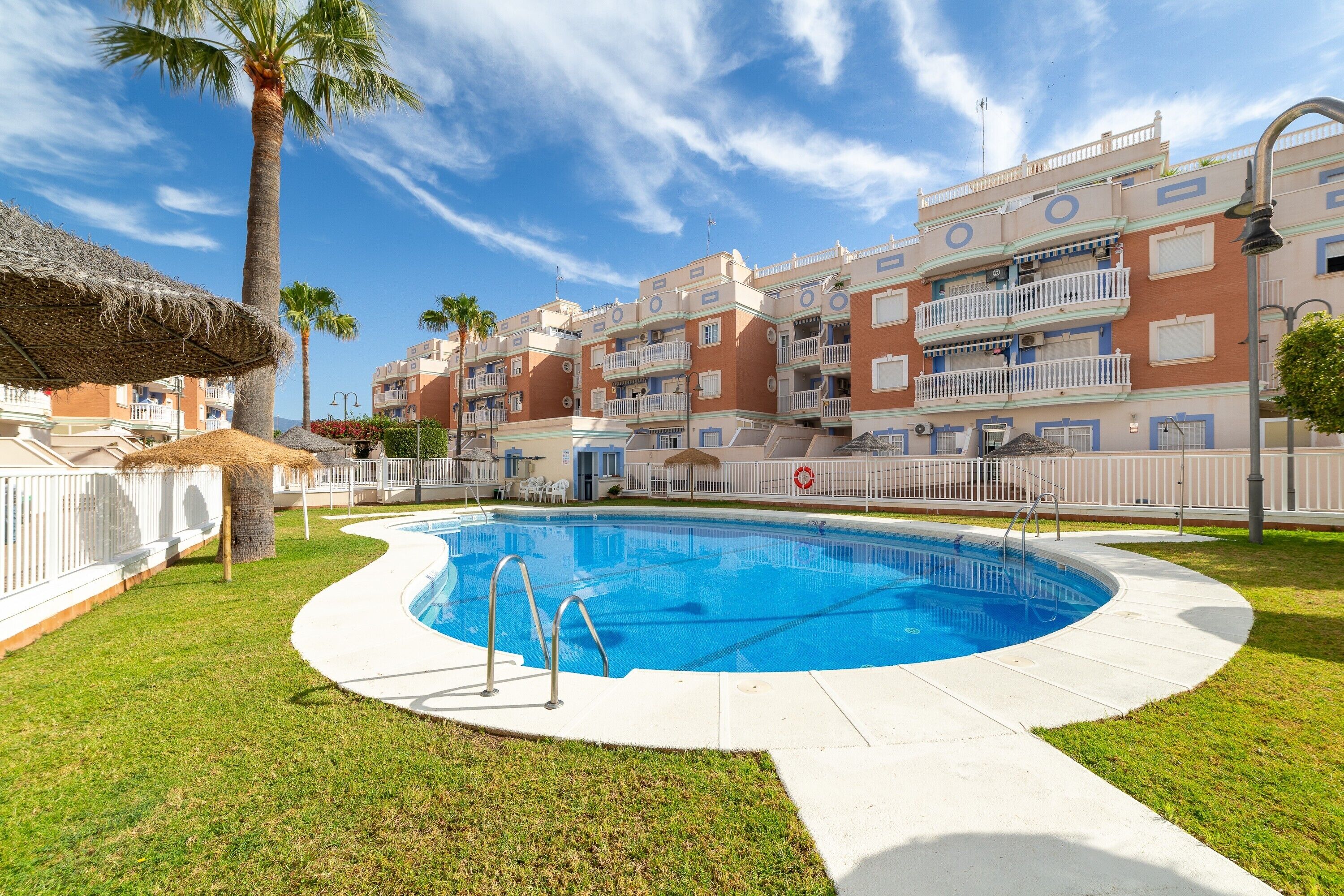 Una piscina rodeada por una valla blanca, con una palmera y un edificio con balcones al fondo.