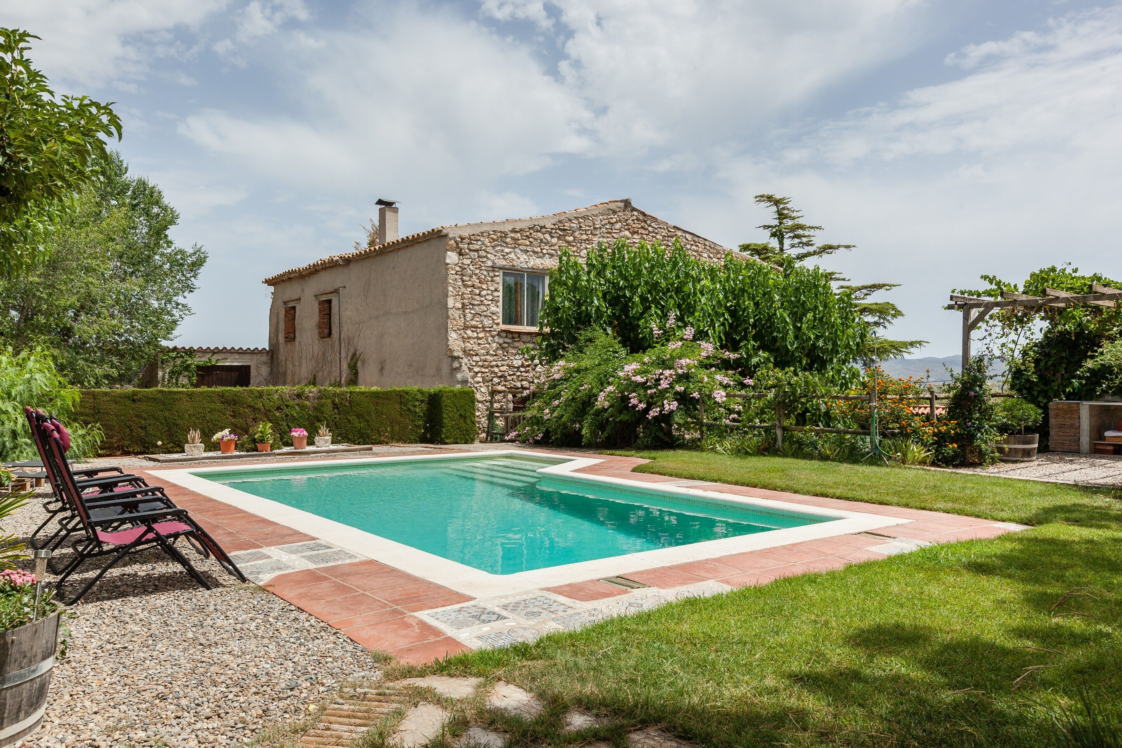 A stone house with a swimming pool, surrounded by greenery and a pergola.