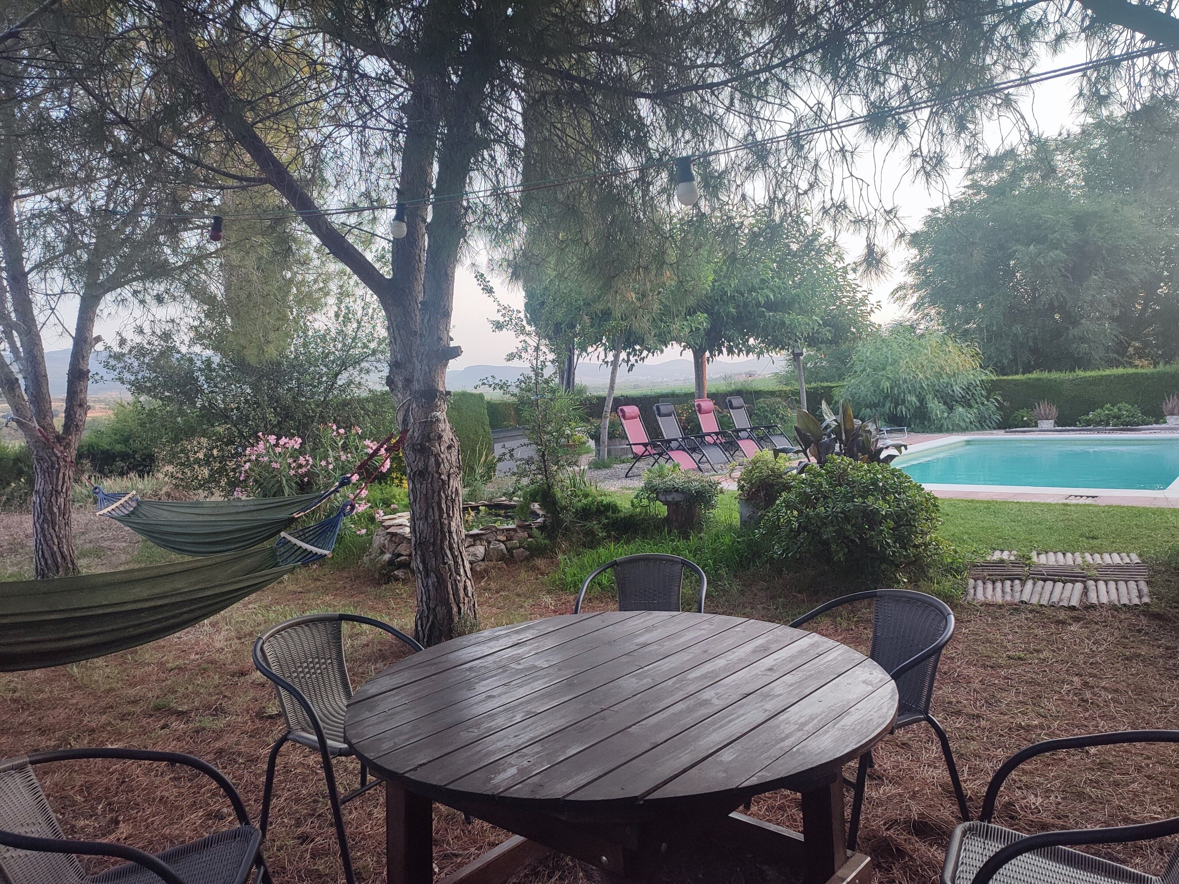 Mesa de madeira com cadeiras, uma rede e uma piscina em um jardim.