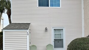 Une maison à deux étages avec un porche à l’avant, deux fauteuils Adirondack verts et un petit abri de jardin.