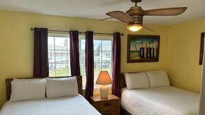 Une chambre d’hôtel avec deux lits, un ventilateur de plafond, une fenêtre avec des rideaux, un tableau et une lampe.